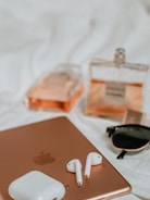 A scene of modern accessories and technology items including a pair of wireless earbuds, a tablet, sunglasses, a charging case, and two bottles of perfume. The items are placed on a soft, white surface. The perfumes have a peachy tint and the tablet is a gold color.