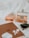 A scene of modern accessories and technology items including a pair of wireless earbuds, a tablet, sunglasses, a charging case, and two bottles of perfume. The items are placed on a soft, white surface. The perfumes have a peachy tint and the tablet is a gold color.