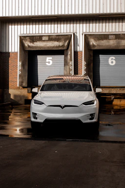 A white electric car is parked in front of industrial garage doors numbered 5 and 6. The car is on a wet pavement, reflecting light from its surroundings. The background consists of a combination of brick and metal siding.