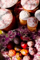 Several baked goods topped with white icing and colorful sprinkles are placed on floral-patterned napkins. Below them, a collection of vibrantly colored and decorated eggs is arranged on similar floral napkins.