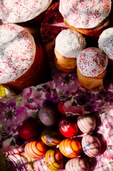 Several baked goods topped with white icing and colorful sprinkles are placed on floral-patterned napkins. Below them, a collection of vibrantly colored and decorated eggs is arranged on similar floral napkins.