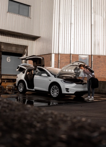 A person stands next to a white Tesla with the hood open in an industrial setting. The car's gull-wing door is raised, and another person sits in the driver's seat. The building in the background has metal siding and a brick base.