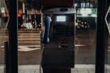A dimly lit, old-fashioned payphone booth located on a city street at night. The phone is blue, with buttons visible on the metallic surface. The street outside is blurred, hinting at a bustling urban environment with some neon signage visible in the background.