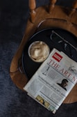 A wooden chair with a round seat is holding a small table tray that contains a cup of coffee with a frothy top, a pair of eyeglasses, and a folded newspaper featuring an article title about archaeology. The scene has a casual, relaxed setup.