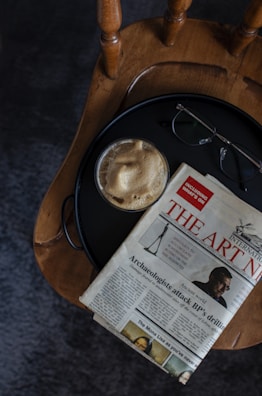 A wooden chair with a round seat is holding a small table tray that contains a cup of coffee with a frothy top, a pair of eyeglasses, and a folded newspaper featuring an article title about archaeology. The scene has a casual, relaxed setup.