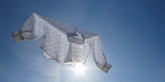 A white blouse with polka dots is suspended in the air against a backdrop of a bright blue sky. The sunlight peeks through the translucent fabric, creating a halo effect around the garment.