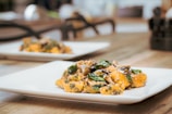 A close-up view of a plate of gourmet food, featuring gnocchi with sautéed mushrooms and spinach, sprinkled with grated cheese. The presentation is elegant, set on a wooden table with a blurred background and natural lighting.