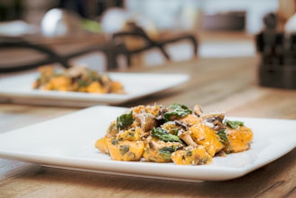 A close-up view of a plate of gourmet food, featuring gnocchi with sautéed mushrooms and spinach, sprinkled with grated cheese. The presentation is elegant, set on a wooden table with a blurred background and natural lighting.