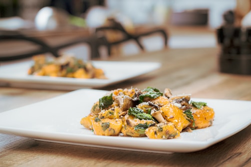A close-up view of a plate of gourmet food, featuring gnocchi with sautéed mushrooms and spinach, sprinkled with grated cheese. The presentation is elegant, set on a wooden table with a blurred background and natural lighting.