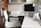 A modern home office setup featuring a sleek white desk with an iMac computer and a white printer. The workspace includes a white leather office chair, a desk lamp, various office supplies, and neatly organized shelves with books and decorative boxes in the background. The setting is well-lit with natural light coming from a large window.