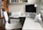 A modern home office setup featuring a sleek white desk with an iMac computer and a white printer. The workspace includes a white leather office chair, a desk lamp, various office supplies, and neatly organized shelves with books and decorative boxes in the background. The setting is well-lit with natural light coming from a large window.