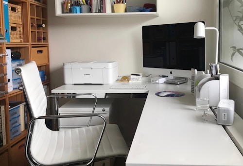A modern home office setup featuring a sleek white desk with an iMac computer and a white printer. The workspace includes a white leather office chair, a desk lamp, various office supplies, and neatly organized shelves with books and decorative boxes in the background. The setting is well-lit with natural light coming from a large window.