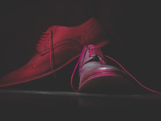 A pair of fashionable leather dress shoes with intricate detailing and brogue patterns is placed against a dark background. The shoes are illuminated with a soft light creating a dramatic shadow effect, highlighting their polished surface and craftsmanship.