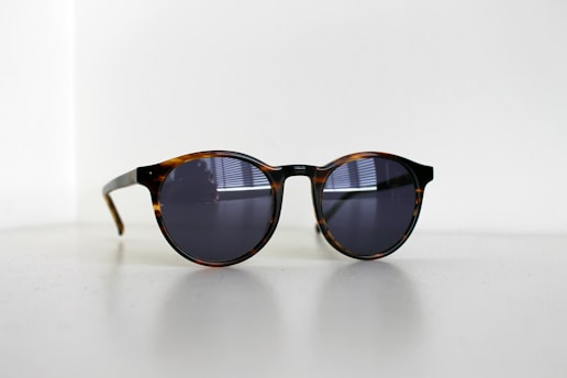 A pair of round sunglasses with dark lenses and a tortoiseshell frame sits on a light surface. Subtle reflections are visible on the lenses, suggesting a nearby window. The lighting in the image is soft, creating gentle shadows.