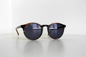A pair of round sunglasses with dark lenses and a tortoiseshell frame sits on a light surface. Subtle reflections are visible on the lenses, suggesting a nearby window. The lighting in the image is soft, creating gentle shadows.