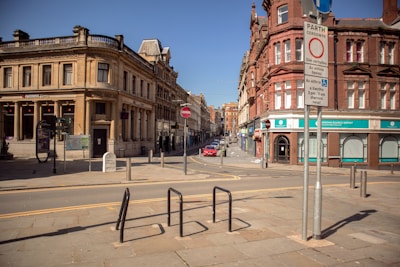 A quiet urban street featuring historic buildings with ornate architecture, including a large NatWest bank with classical columns on the left. The street is lined with bollards and road signs, and a red car is parked further down the street. The atmosphere is calm and the sky is clear.
