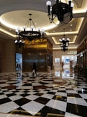 An elegant interior space with a high ceiling adorned with large, ornate chandeliers. The floor features a striking geometric black and white pattern. A person is walking across the reflective surface, carrying a shopping bag. In the background, there are shops with various displays.