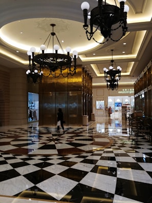 An elegant interior space with a high ceiling adorned with large, ornate chandeliers. The floor features a striking geometric black and white pattern. A person is walking across the reflective surface, carrying a shopping bag. In the background, there are shops with various displays.