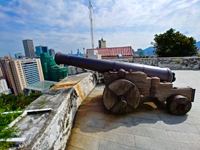 A historical cannon mounted on wooden wheels is placed on a stone platform with a cityscape in the background. The cannon is old and weathered, suggesting historical significance or preservation. Skyscrapers and buildings form the backdrop under a partly cloudy sky, with lush greenery also visible.