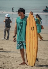 A man wearing a black cap, teal tank top, and patterned shorts stands on a sandy beach, holding a wooden surfboard. People walk in the background near the shoreline and a person wearing a white hat is taking a photo.