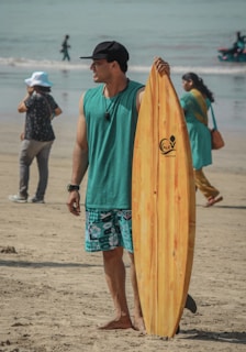 A man wearing a black cap, teal tank top, and patterned shorts stands on a sandy beach, holding a wooden surfboard. People walk in the background near the shoreline and a person wearing a white hat is taking a photo.