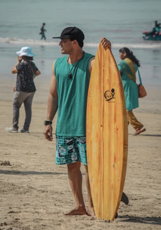A man wearing a black cap, teal tank top, and patterned shorts stands on a sandy beach, holding a wooden surfboard. People walk in the background near the shoreline and a person wearing a white hat is taking a photo.