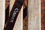 A black camera strap with the brand name embroidered in white, set against a textured wooden surface. The wood appears weathered with a natural, light brown hue. The overall setting suggests an outdoor environment.