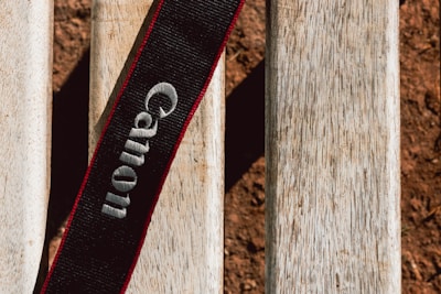 A black camera strap with the brand name embroidered in white, set against a textured wooden surface. The wood appears weathered with a natural, light brown hue. The overall setting suggests an outdoor environment.
