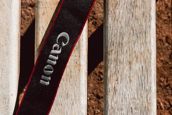 A black camera strap with the brand name embroidered in white, set against a textured wooden surface. The wood appears weathered with a natural, light brown hue. The overall setting suggests an outdoor environment.