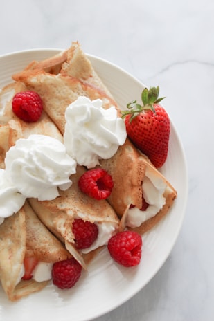 A plate of crepes filled with cream, garnished with whipped cream, fresh raspberries, and a whole strawberry.