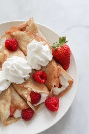A plate of crepes filled with cream, garnished with whipped cream, fresh raspberries, and a whole strawberry.