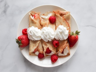 A white plate holds four crepes filled with whipped cream and garnished with fresh strawberries and raspberries. Each crepe is topped with a dollop of whipped cream, and additional berries are placed around the crepes for decoration. The backdrop is a light, marble-textured surface.
