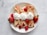A white plate holds four crepes filled with whipped cream and garnished with fresh strawberries and raspberries. Each crepe is topped with a dollop of whipped cream, and additional berries are placed around the crepes for decoration. The backdrop is a light, marble-textured surface.