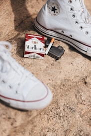 A pack of Marlboro cigarettes, an open lighter, and a half-smoked cigarette are placed on a rough, sandy surface. A pair of white Converse All Star high-top shoes are also visible, suggesting a casual, possibly rebellious setting.