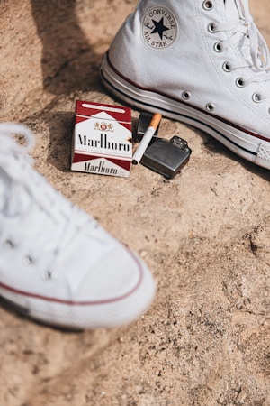 A pack of Marlboro cigarettes, an open lighter, and a half-smoked cigarette are placed on a rough, sandy surface. A pair of white Converse All Star high-top shoes are also visible, suggesting a casual, possibly rebellious setting.