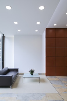 A modern interior with minimalist design featuring a sleek black couch next to a large window. A glass coffee table with a small plant sits on a light-colored rug over a tiled floor. The space is illuminated by ceiling lights and is partially enclosed by a wall with a reddish-brown panel design.