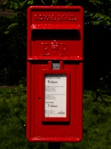 A red Royal Mail postbox with the emblem 'E II R' prominently displayed. Instructions for mail collection times are visible on the front panel. The box is surrounded by a backdrop of green foliage and grass.