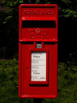 A red Royal Mail postbox with the emblem 'E II R' prominently displayed. Instructions for mail collection times are visible on the front panel. The box is surrounded by a backdrop of green foliage and grass.
