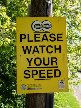 A bright yellow sign is mounted on a wooden pole with the text 'PLEASE WATCH YOUR SPEED' in bold black letters. Above the text is a logo with the words 'Speed Watch' and below is a reference to Oxfordshire County Council. Surrounding the sign are lush green leaves from nearby trees.