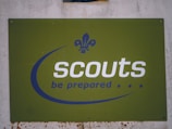 A green sign with the word 'scouts' written in bold white letters and the motto 'be prepared' below it in blue. The sign features a fleur-de-lis logo in blue, positioned above the text. The background is slightly worn, indicating some weathering.