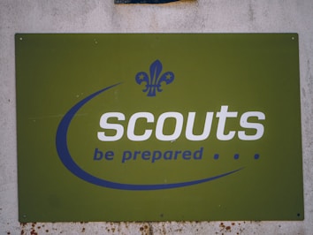 A green sign with the word 'scouts' written in bold white letters and the motto 'be prepared' below it in blue. The sign features a fleur-de-lis logo in blue, positioned above the text. The background is slightly worn, indicating some weathering.