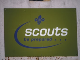 A green sign with the word 'scouts' written in bold white letters and the motto 'be prepared' below it in blue. The sign features a fleur-de-lis logo in blue, positioned above the text. The background is slightly worn, indicating some weathering.