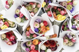 Several bouquets of colorful flowers are arranged in white paper wraps. The flowers include roses, ranunculus, and peonies in a variety of vibrant hues such as red, pink, purple, orange, and yellow. The bouquets are laid out in a symmetrical pattern on a surface.