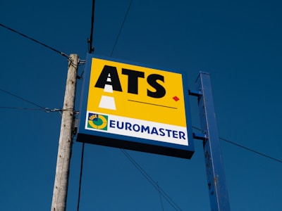 A signboard mounted on a metal post and wooden pole against a clear blue sky. The sign features the large letters 'ATS' on a yellow background with a stylized road graphic. Below, it reads 'EUROMASTER' in blue text next to a circular logo with a yellow and green color scheme.