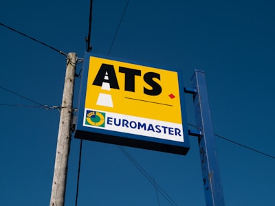 A signboard mounted on a metal post and wooden pole against a clear blue sky. The sign features the large letters 'ATS' on a yellow background with a stylized road graphic. Below, it reads 'EUROMASTER' in blue text next to a circular logo with a yellow and green color scheme.