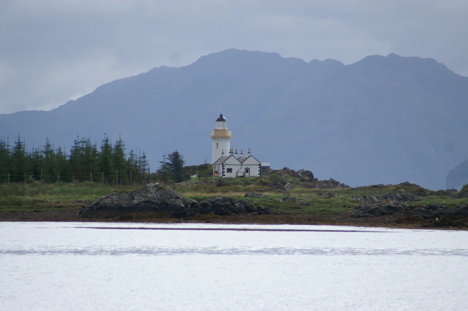The image depicts a serene coastal scene with a white lighthouse standing tall on a rocky outcropping, overlooking a body of water. The lighthouse is surrounded by lush greenery and trees, creating a picturesque backdrop for the structure. In the distance, majestic mountains rise against an overcast sky, adding depth to the landscape.