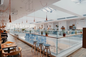 A modern open-plan cafe with wooden floors and sleek pendant lights. The space features a glass railing and several tables with chairs. Potted plants add a touch of greenery, and the area is filled with natural light streaming from large windows and skylights.