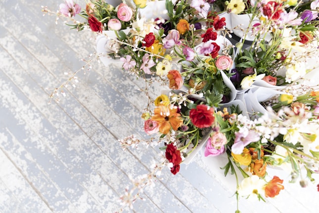 A collection of vibrant, multi-colored flowers arranged in small bouquets with paper wraps. The flowers include varieties with delicate petals and vivid hues, such as reds, pinks, yellows, and greens. They are placed on a weathered wooden surface, which has a rustic and textured appearance.