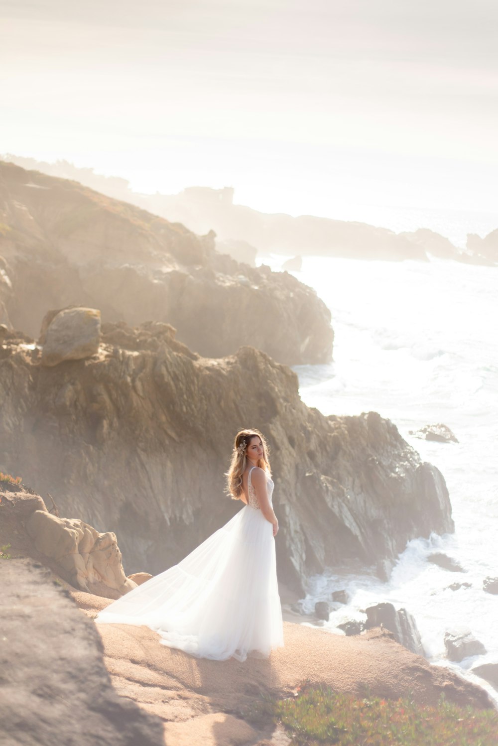Frau im weißen Hochzeitskleid, die tagsüber auf einer Felsformation in der Nähe eines Gewässers steht