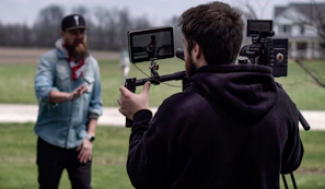 Two men are outside in a somewhat rural setting, possibly a park or a field. One man, wearing a blue shirt and a black cap, appears to be talking or gesturing towards the camera. The other man is operating camera equipment, which includes a monitor showing the man in front of the camera. The cameraman is wearing a black hoodie.